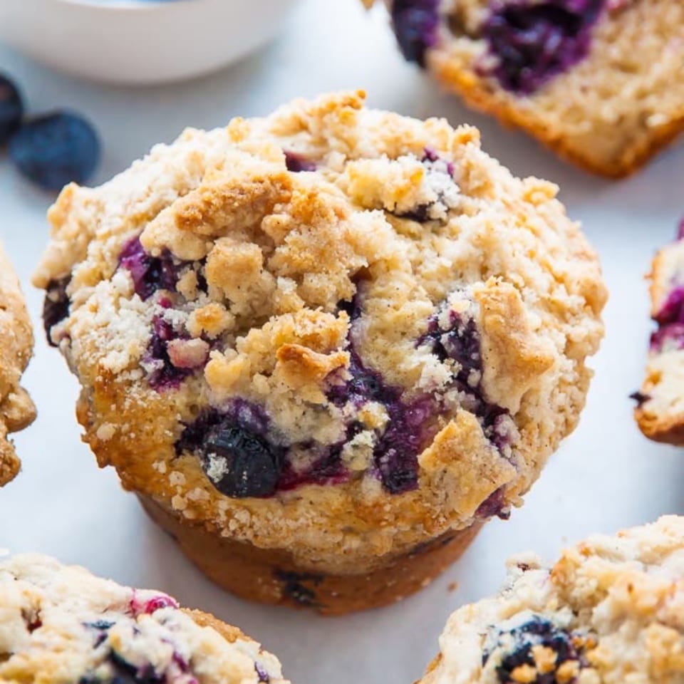 Blueberry muffin topped with golden crumb streusel, surrounded by additional muffins on a baking tray.