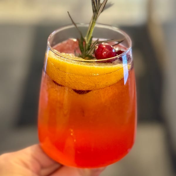 A vibrant orange and red cocktail, garnished with orange slices, cranberries, and a sprig of rosemary.