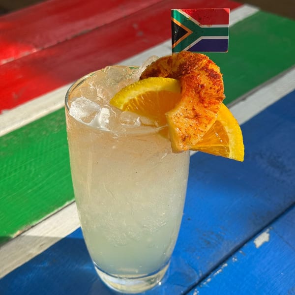 Iced cocktail with orange slices and a South African flag pick on a colorful striped table.