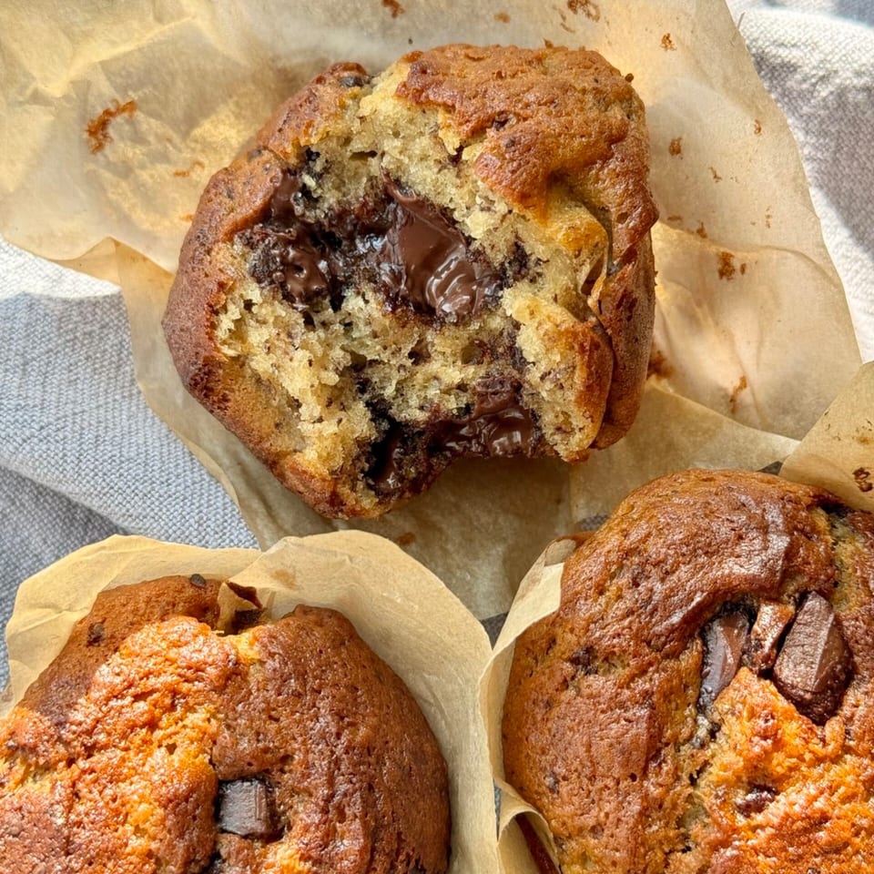 Three golden-brown chocolate chunk muffins arranged on parchment paper, with one broken open to reveal a gooey chocolate center.