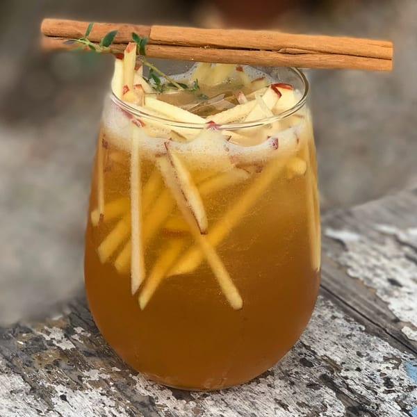A glass of amber-colored drink with apple slices, a cinnamon stick, and a sprig of thyme.