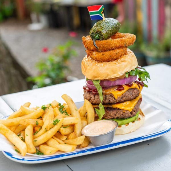 A towering double cheeseburger with onion rings, a jalapeño, and a South African flag, served with fries.