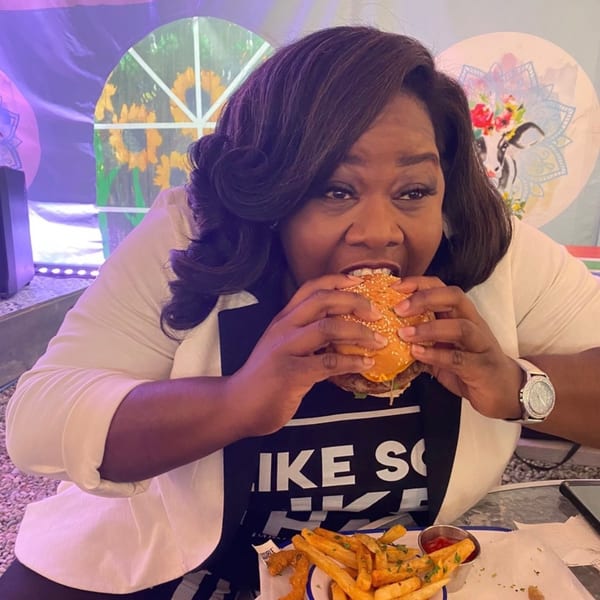 Woman in a white blazer taking a large bite out of a burger, with fries on the table.
