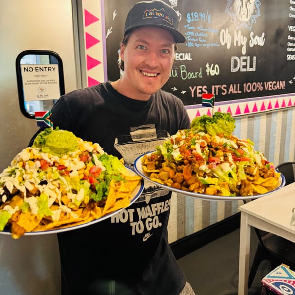 A smiling man in a cap holds two large plates of vegan nachos topped with guacamole and salsa.