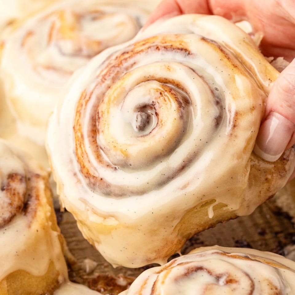 Close-up of a glazed cinnamon roll with visible swirls, held by hand among other pastries.