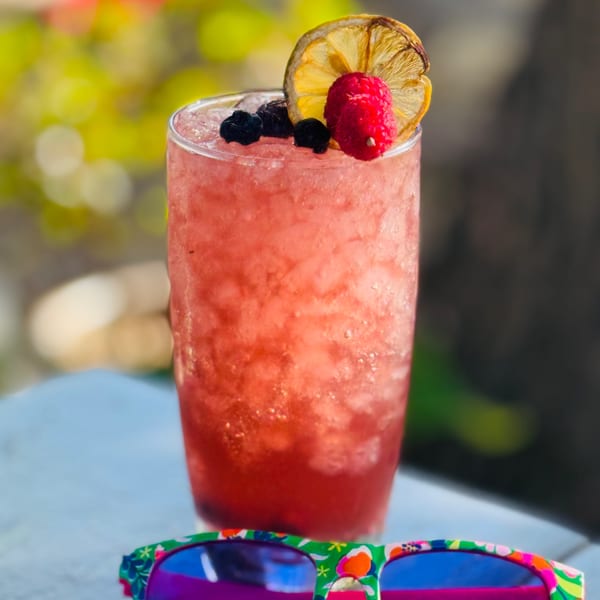 A vibrant red cocktail with berries and a dried lemon slice, next to colorful sunglasses.