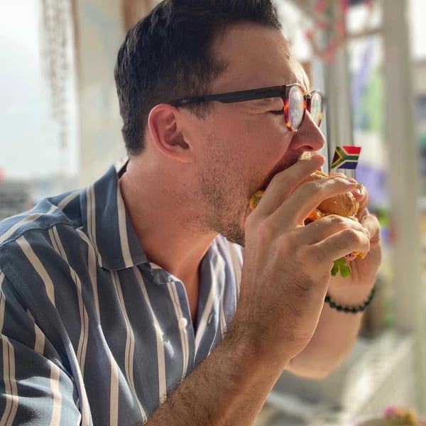 Man in glasses and a striped shirt taking a large bite out of a burger with a South African flag pick.
